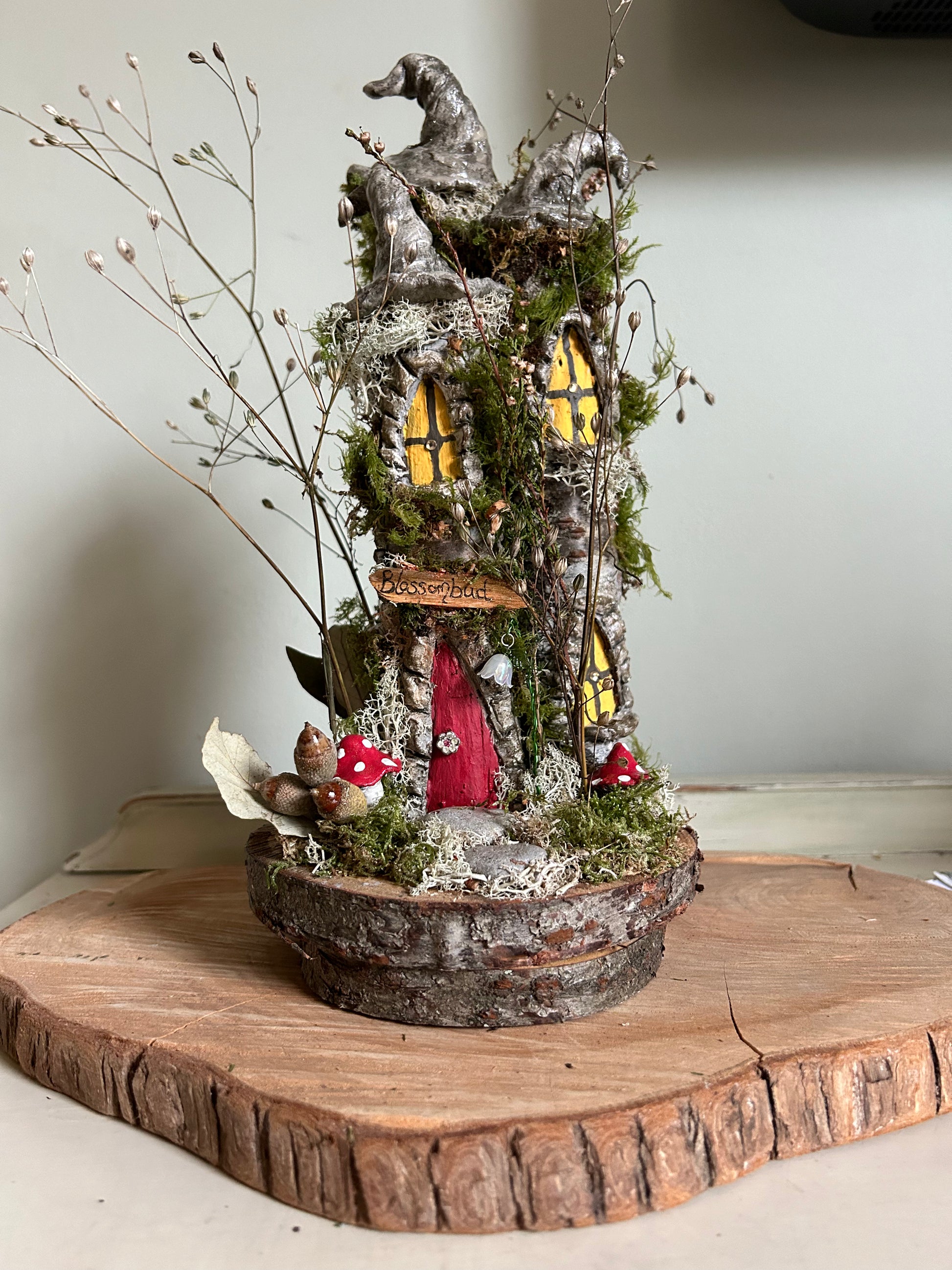 A decorative wooden house with a bark roof, adorned with flowers, toadstools, and a name plaque, crafted from natural materials and designed to resemble a fae elemental dwelling.